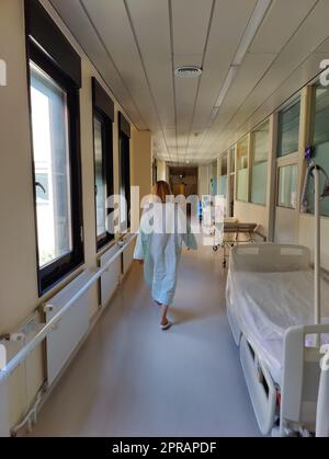 Paziente femminile che indossa l'abito dell'ospedale che cammina nel lungo corridoio vuoto dell'ospedale. Foto Stock