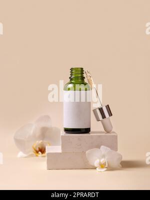 Bottiglia contagocce verde aperta vicino a fiori di orchidea bianchi su pietra, etichetta cosmetica Mockup Foto Stock