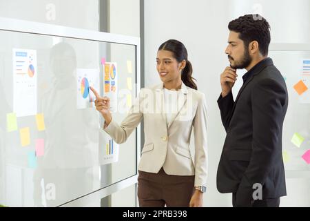 Giovane donna d'affari asiatica in tuta marrone che punta il dito sul grafico a torta per l'obiettivo aziendale, presentata all'investitore. Sul pannello di vetro erano presenti una carta per banconote e un documento. Foto Stock