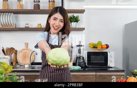 Giovane casalinga asiatica vestita in un grembiule che offre lattuga fresca nella parte anteriore con entrambe le mani. Atmosfera mattutina in una cucina moderna. Foto Stock