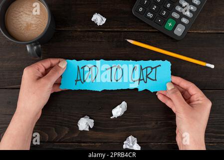 Testo calligrafia Aggiungi al carrello. Approccio aziendale acquisto online e-commerce tecnologie moderne per fare acquisti a mani femminili che tengono carta bianca e cancelleria su un tavolo di legno. Foto Stock