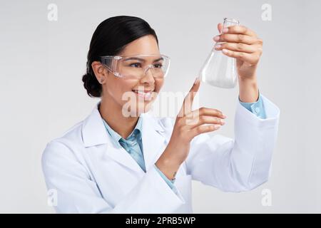 Questa è la reazione che sta cercando. Una giovane scienziata femminile attraente che esamina un becher riempito di liquido in studio su uno sfondo grigio. Foto Stock