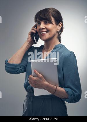 Sempre pronti e in attesa della prossima opportunità. Foto in studio di una donna anziana che parla su un cellulare mentre tiene in mano un tablet digitale su uno sfondo grigio. Foto Stock