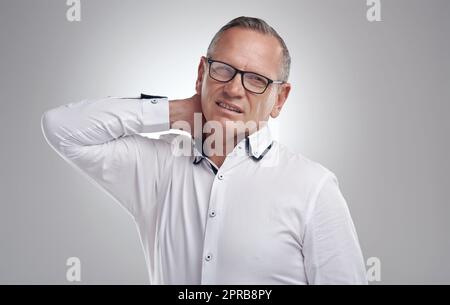 Im ottenere un po 'vecchio per questo. Un bel uomo d'affari maturo in piedi da solo su uno sfondo grigio in studio e che soffre di mal di collo. Foto Stock