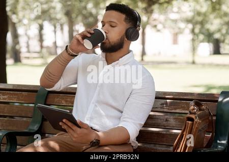 Goditi una breve pausa caffè all'aria aperta. un giovane uomo d'affari che indossa delle cuffie e usa un tablet digitale mentre è seduto su una panchina all'aperto. Foto Stock