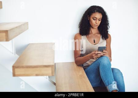 Donna che si rilassa, guarda e scrive al telefono seduta su una scala di legno. Lettura femminile afroamericana attraente, informale e sicura su cellulare. Fare una pausa e recuperare i contatti sui social media Foto Stock