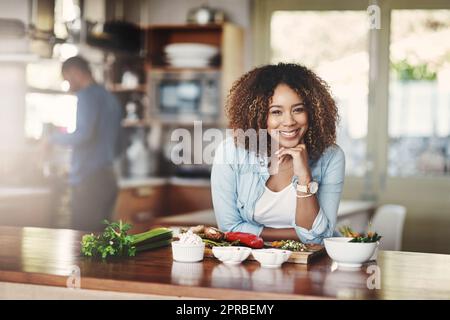 Ritratto di una giovane donna felice, sana e spensierata che prepara un pasto sano a casa con il marito sullo sfondo. La moglie nera prepara un'insalata vegetariana biologica per pranzo in cucina Foto Stock