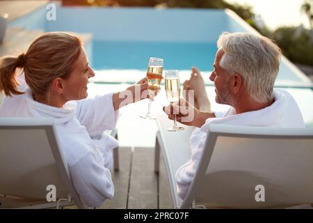 Celebrare il loro amore con stile. Ripresa di una coppia matura che beve champagne mentre si rilassa sulle sedie a sdraio al tramonto. Foto Stock