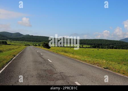 Una strada asfaltata a due corsie con crepe passa attraverso una valle pianeggiante con cespugli sparsi verso alte colline in una giornata di sole estate. Khakassia, Siberia, Russia. Foto Stock