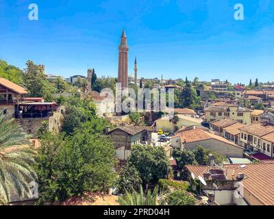 piccole case con tetti di tegole sullo sfondo di palme verdi. al centro della piazza si trova un'alta torre con una guglia. Foto Stock