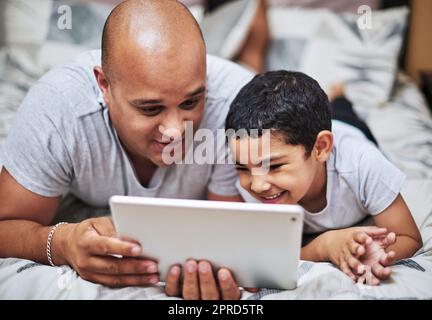 Quindi questo è lo spettacolo che ti piace tanto. un giovane allegro e suo figlio che guardano video su un tablet digitale mentre si stendono a casa in un letto durante il giorno. Foto Stock