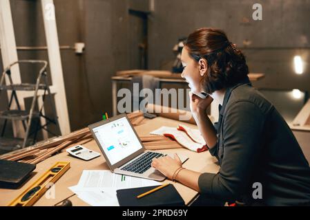 Ho alcune cose da cui non scegliere. Un carpentiere femminile attraente che lavora sul suo laptop mentre parla sul suo cellulare all'interno di un laboratorio. Foto Stock