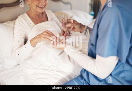 Una dose giornaliera di salute che arriva a destra. Un'infermiera che dà ad una donna anziana il suo farmaco da prendere con un bicchiere d'acqua nel letto. Foto Stock