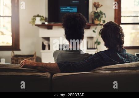 Una coppia che fluisce insieme, rimane insieme. Foto con vista posteriore di una giovane coppia seduta sul divano e guardando la tv insieme a casa. Foto Stock