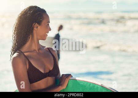 Sorridi e agita: Una donna in spiaggia con la sua tavola da surf mentre un surfista maschile si alza sullo sfondo. Foto Stock