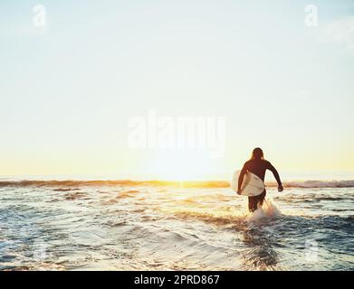 Sia di destra indietro - devo andare prendere alcune onde. Dietro l'inquadratura di un giovane che corre verso il mare mentre porta una tavola da surf in spiaggia. Foto Stock