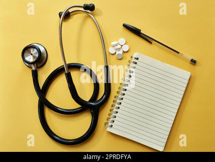 Proprio ciò che il medico ha prescritto. Studio di uno stetoscopio, blocco note, penna e pillole su sfondo giallo. Foto Stock