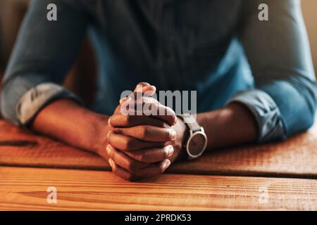 Uomo tranquillo, calmo e spirituale che prega seduto con le mani ripiegate da solo a casa. Primo piano pregando, speranzoso e religioso, cristiano maschio che dice una preghiera quotidiana al mattino a un tavolo Foto Stock