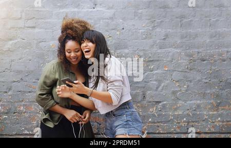 I social media ci fanno sempre ridere ad alta voce. Due giovani donne allegre che usano un cellulare insieme mentre si posano all'aperto contro un muro di mattoni grigi. Foto Stock