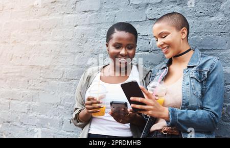 Connettiti ovunque e in qualsiasi momento. Due giovani donne che stanno accanto a un edificio sorridendo e leggendo messaggi di testo mentre tengono le loro bevande fresche. Foto Stock