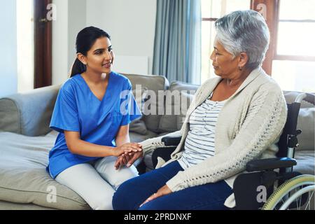 Continua ad essere ottimista e andrà tutto bene. una donna anziana dall'aspetto preoccupata seduta su una sedia a rotelle mentre un'infermiera le tiene la mano per sostenerla all'interno di casa durante il giorno. Foto Stock