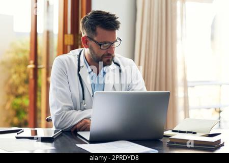 Scrivere un rapporto sul paziente è appena visto. Un bel medico maschile che lavora sul suo computer portatile mentre si siede in ufficio. Foto Stock