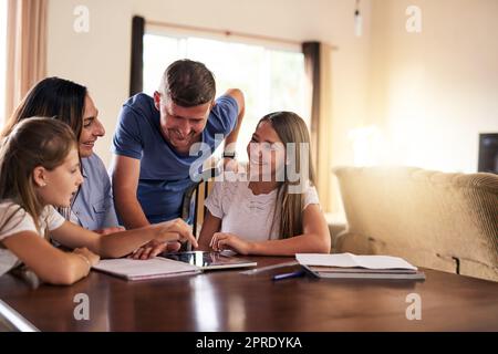 Grazie per l'aiuto di mamma e papà. Due ragazze allegre fare i compiti a casa intorno a un tavolo, mentre ottenere aiuto dai loro genitori a casa durante il giorno. Foto Stock