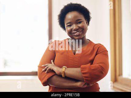 Ritratto di una donna d'affari afro-americana sorridente e informale, in piedi sicura di sé con le braccia incrociate in un ufficio moderno. Designer professionista allegra che lavora in una società di startup Foto Stock