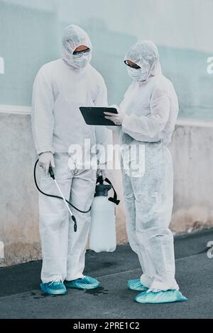 Operatori sanitari che puliscono fuori da un edificio utilizzando un elenco per seguire le istruzioni sulla sicurezza dei rischi biologici durante la covid. Ricercatori medici che indossano tute di nocciolamento sanificazione all'aperto per prevenire le infezioni Foto Stock