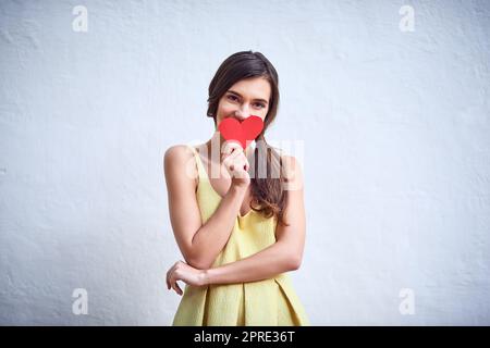 A chi potrebbe pensare? Foto in studio di una giovane donna allegra che tiene in mano un pezzo di carta a forma di cuore mentre si trova su uno sfondo grigio. Foto Stock