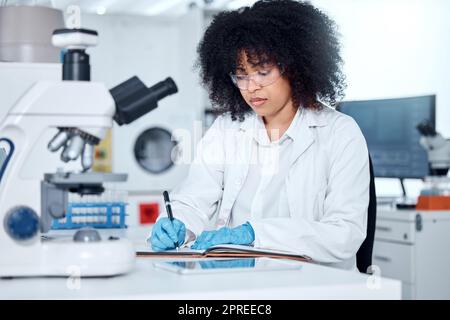 Uno scienziato di razza mista con capelli ricci che indossano l'apparecchiatura di sicurezza che scrive le note e che analizza i campioni medici di prova con il microscopio in laboratorio. Donna che fa Foto Stock