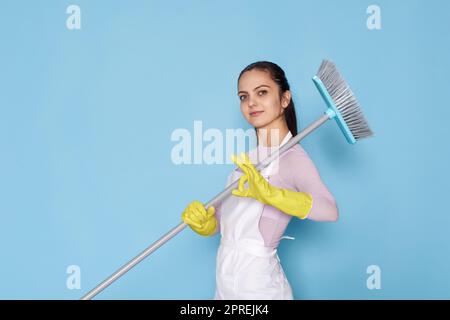 donna caucasica in guanti di gomma e grembiule più pulito che tiene la scopa e mostra ok gestureon sfondo blu. Foto Stock