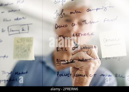 Pensateci, pianificatelo e poi eseguirlo. Un uomo d'affari senior che scrive le idee con un pennarello su una parete di vetro in ufficio. Foto Stock