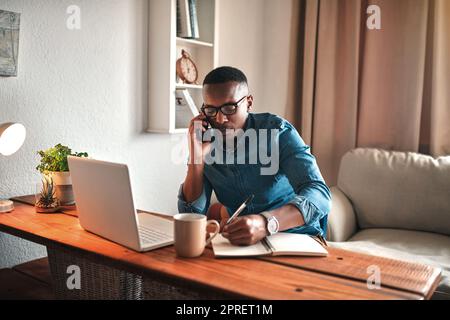 Giovane imprenditore al telefono mentre prende appunti alla sua scrivania. Uomo professionista che lavora da casa con la tecnologia. Un uomo afro-americano ha una riunione d'affari con una chiamata audio. Foto Stock