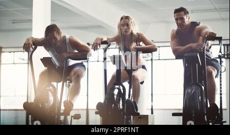 Fare una pausa tra un allenamento e l'altro. Scatto completo di tre giovani seduti su istruttori ellittici mentre si allenano in palestra. Foto Stock