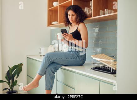 Donna afro al telefono che scrive, invia SMS o legge notizie false online a casa bevendo caffè o tè. Donne alla moda, giovani ed eleganti sedute sul bancone della cucina navigando su Internet e sui social media. Foto Stock