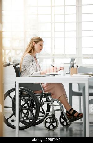 Donna d'affari professionista e disabile in sedia a rotelle che legge documenti, scrive o prende appunti sulla scrivania dell'ufficio seduto accanto a un computer portatile. Imprenditrice donna con disabilità che fa documenti contrattuali con la penna. Foto Stock