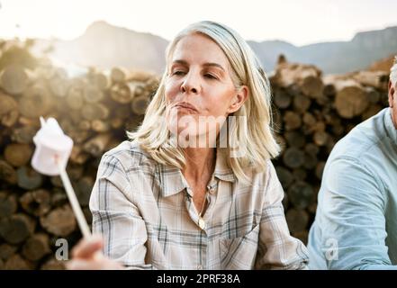 Donna anziana che mangia marshmallow con rammarico, si rilassa accanto al fuoco dopo la sua avventura in montagna nella natura all'aria aperta. Donna che ama il benessere e riposa dopo un'escursione con gli amici Foto Stock