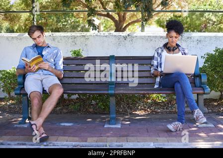 Perso nei nostri mondi separati. due studenti universitari che studiano mentre si siedono su una panchina del campus Foto Stock