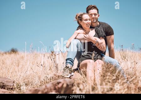 Spensierata, sorridente e rilassata coppia che si unisce, divertendosi e guardando la vista mentre si siede sull'erba in un parco naturale insieme. Fidanzato amorevole, premuroso e romantico e fidanzato che abbraccia Foto Stock