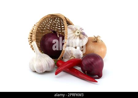 Cipolle rosse, peperoncino rosso e bulbi d'aglio cadono dal cestino di vimini. Spezie e tema aromatizzante ancora vita isolata su sfondo bianco Foto Stock