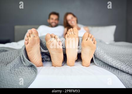 La domenica è il giorno del letto. Una giovane coppia felice che si rilassa a a letto con i piedi che scopano da sotto le lenzuola. Foto Stock