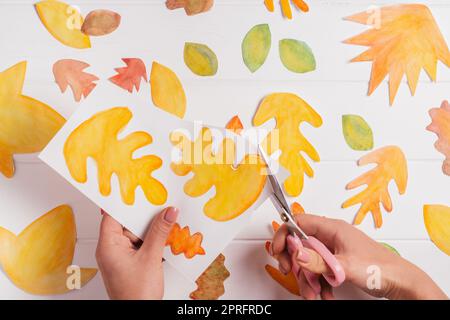Le mani femminili tagliano con forbici disegnate su fogli autunnali di carta bianca di forme, dimensioni e colori diversi. Tagliare le foglie su un tavolo di legno bianco. Foto Stock