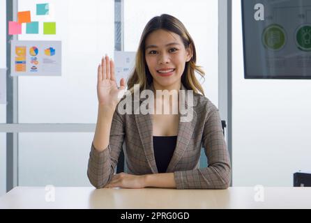 ํYoung impiegata asiatica in un abito a quadri siede e rilassati con il viso sorridente, agitando la mano davanti alla scrivania. C'era una carta per banconote e un documento su una lastra di vetro sullo sfondo. Foto Stock