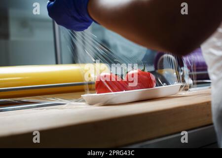 Un lavoratore sconosciuto avvolge i pomodori in pellicola trasparente per alimenti appoggiati su un vassoio di plastica bianco Foto Stock