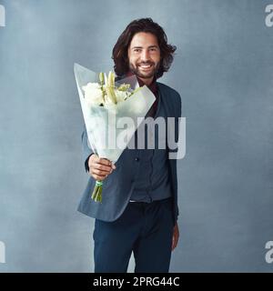 Egli sa come girare il romanticismo. Studio shot di un bel giovane vestito elegantemente con un bouquet di fiori su sfondo grigio. Foto Stock