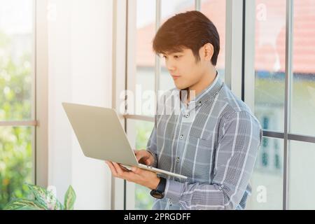 uomo d'affari sorridente e che lavora su un computer portatile stand vicino alla finestra in ufficio Foto Stock