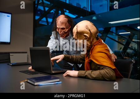Dipendenti Zombie che cercano un notebook e parlano in ufficio notturno. Uomini d'affari mostro non morti che analizzano il risultato del progetto di affari in linea Foto Stock