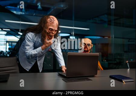 Dipendenti Zombie che cercano un notebook e parlano in ufficio notturno. Uomini d'affari mostro non morti che analizzano il risultato del progetto di affari in linea Foto Stock