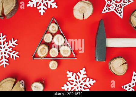 Chiodi lunghi e tagli di legno o lastre di ramo, disposti a forma di albero di Natale con martello e fiocchi di neve in legno su sfondo rosso. Disposizione piatta Foto Stock